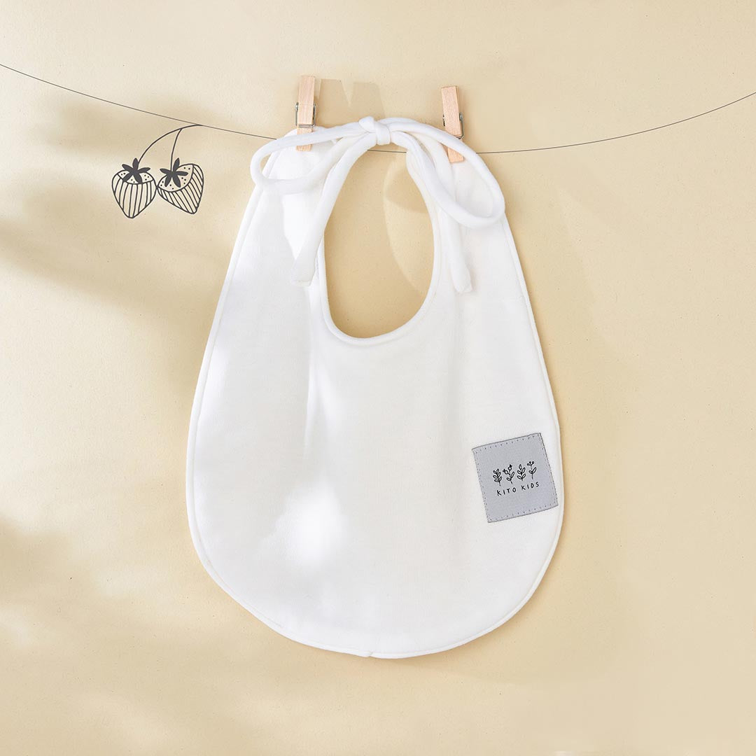 Bavoir de naissance couleur blanc cassé de la marque Kito Kids avec ses attaches blanches nouées en boucle.