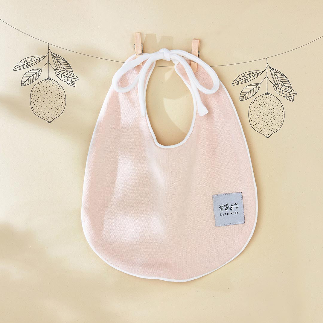 Bavoir de naissance couleur pêche clair de la marque Kito Kids avec des attaches blanches croisées.