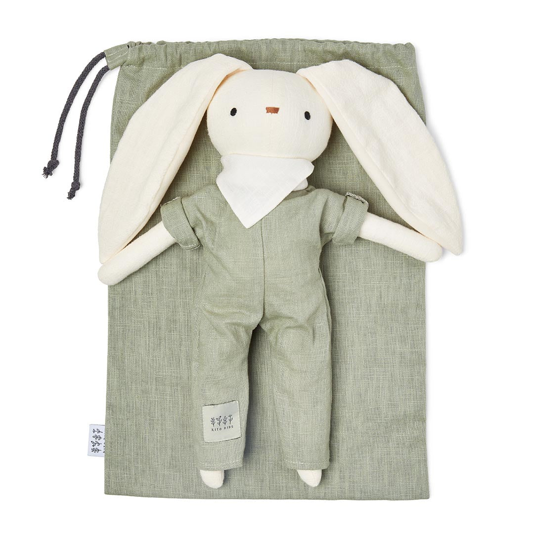 Peluche lapin blanc Kito Kids vêtu d'une combinaison vert sauge avec petit foulard blanc, posé à plat sur son pochon en lin assorti.