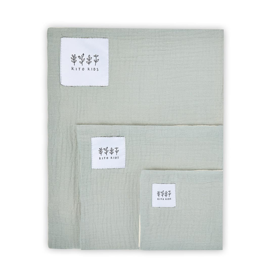 Trois langes en mousseline de coton vert sauge de tailles différentes pliés et superposés sur fond blanc.