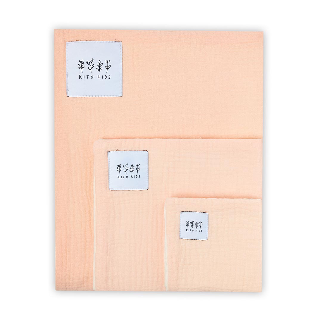 Lot de trois langes en mousseline de coton pêche de tailles différentes, pliés et superposés sur un fond blanc.