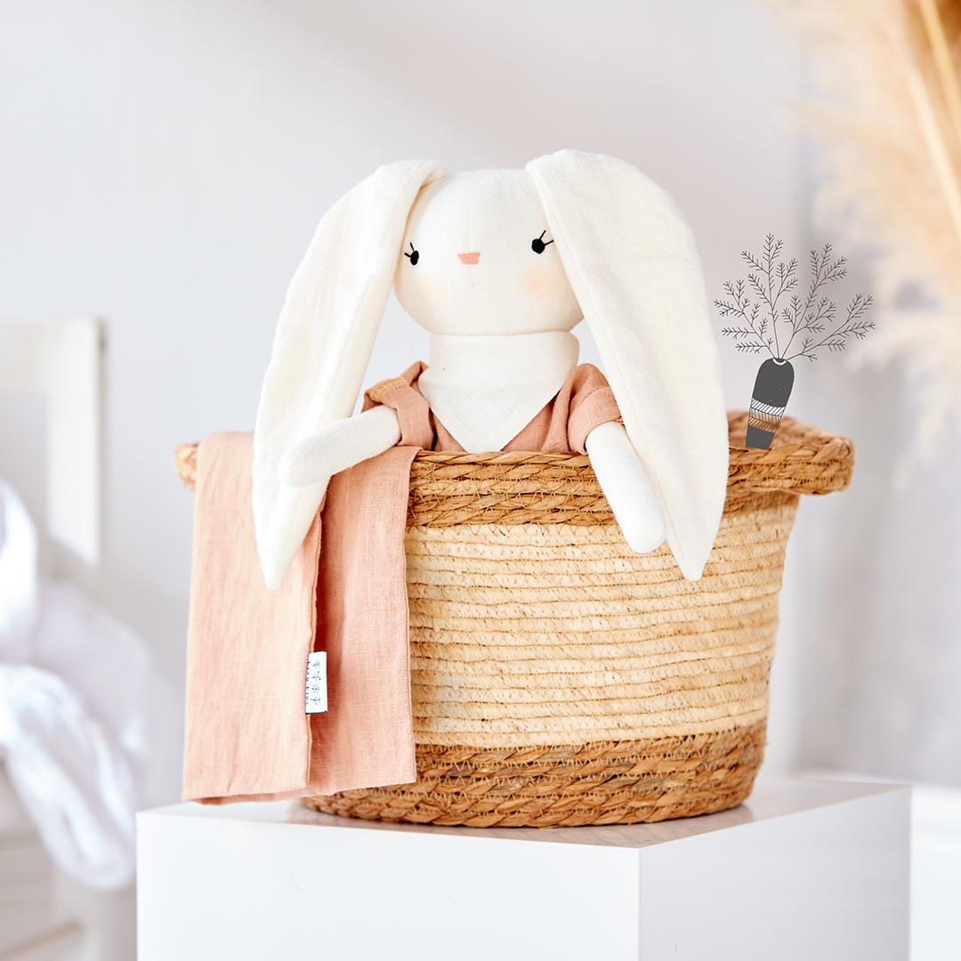 Peluche lapin blanc Kito Kids vêtu d'une combinaison terracotta avec foulard blanc, posé à plat sur son pochon en lin pêche assorti.