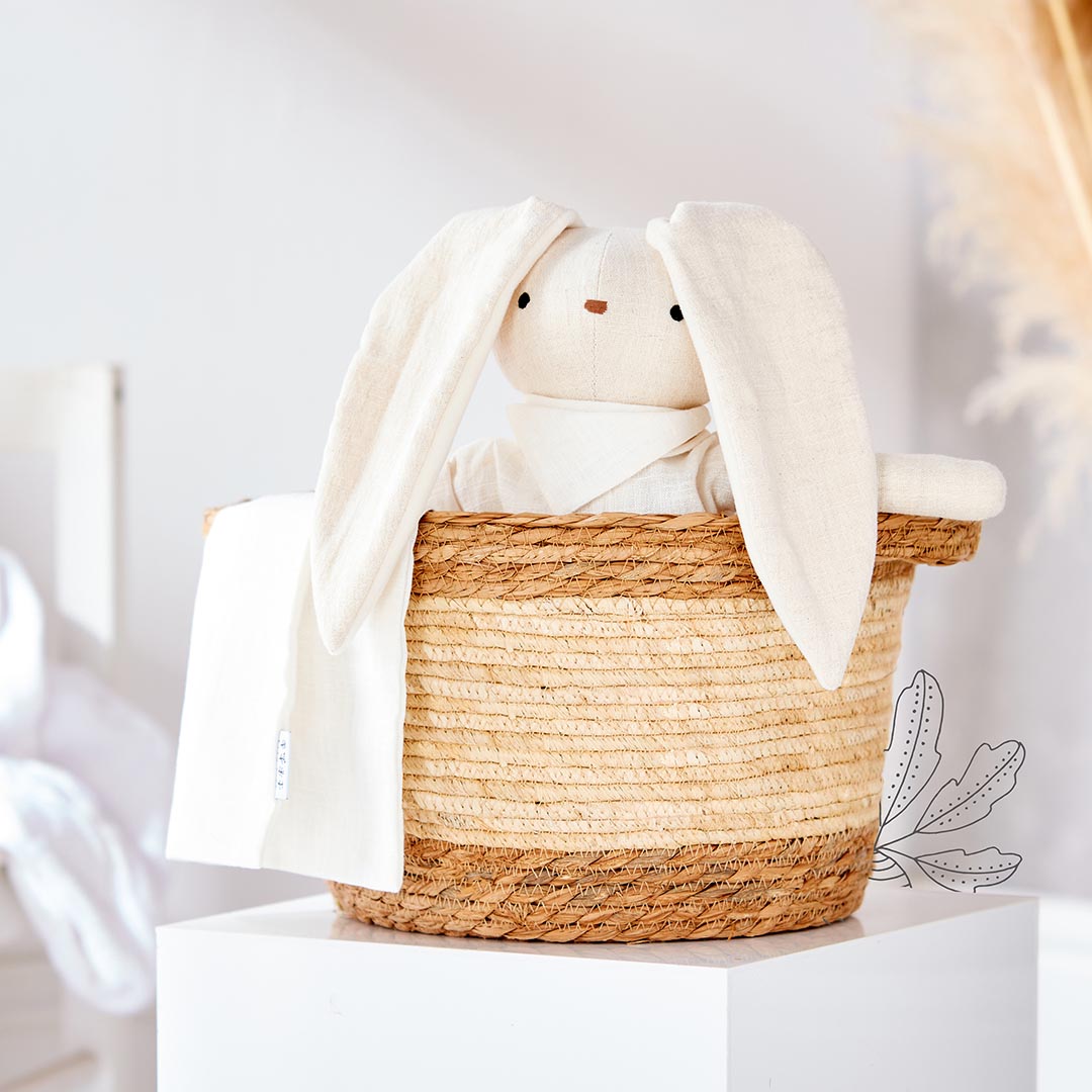 Doudou lapin aux longues oreilles tombantes de couleur beige clair, assis dans un panier tressé avec son lange assorti.