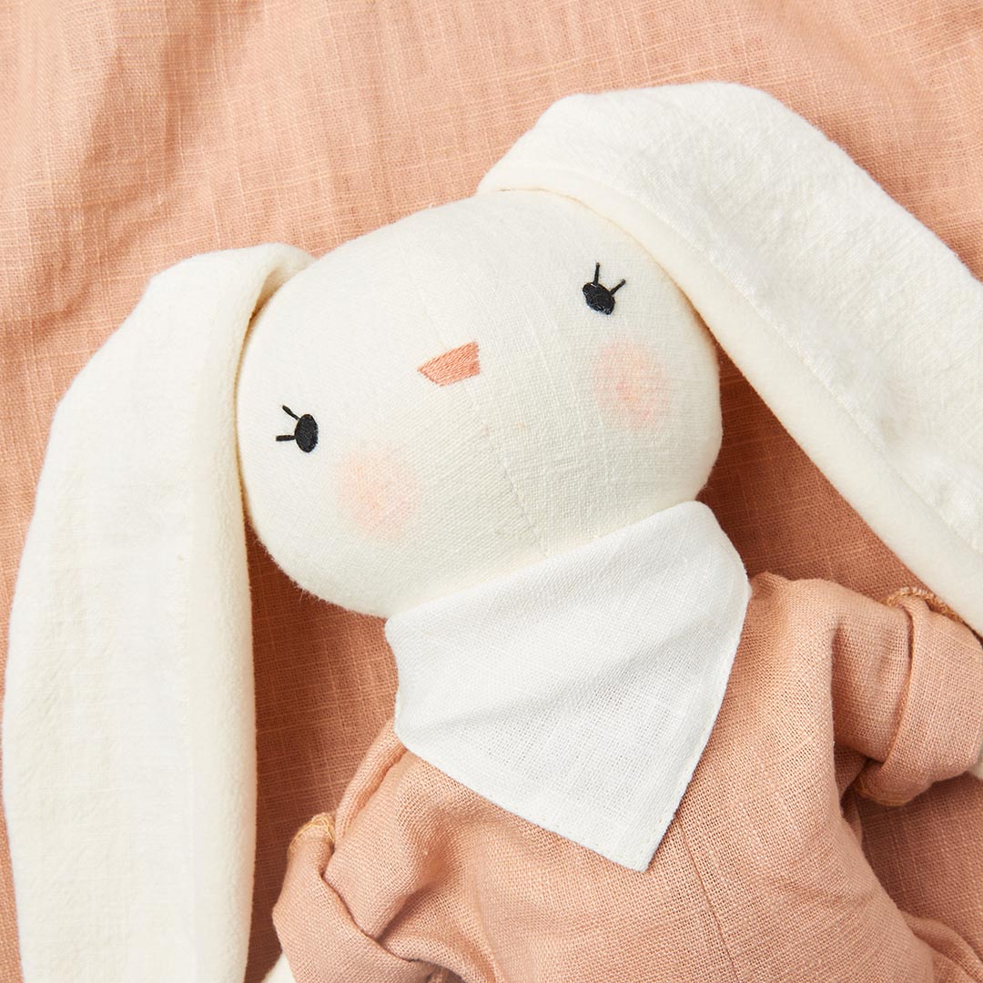 Doudou lapin aux longues oreilles blanches et habit terracotta, assis dans un panier tressé avec un lange pêche suspendu.