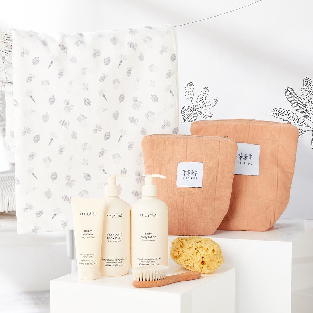 Serviette de bain blanche imprimée pour bébé à côté de deux trousses de toilette matelassées terracotta Kito Kids et de crèmes Mushie.