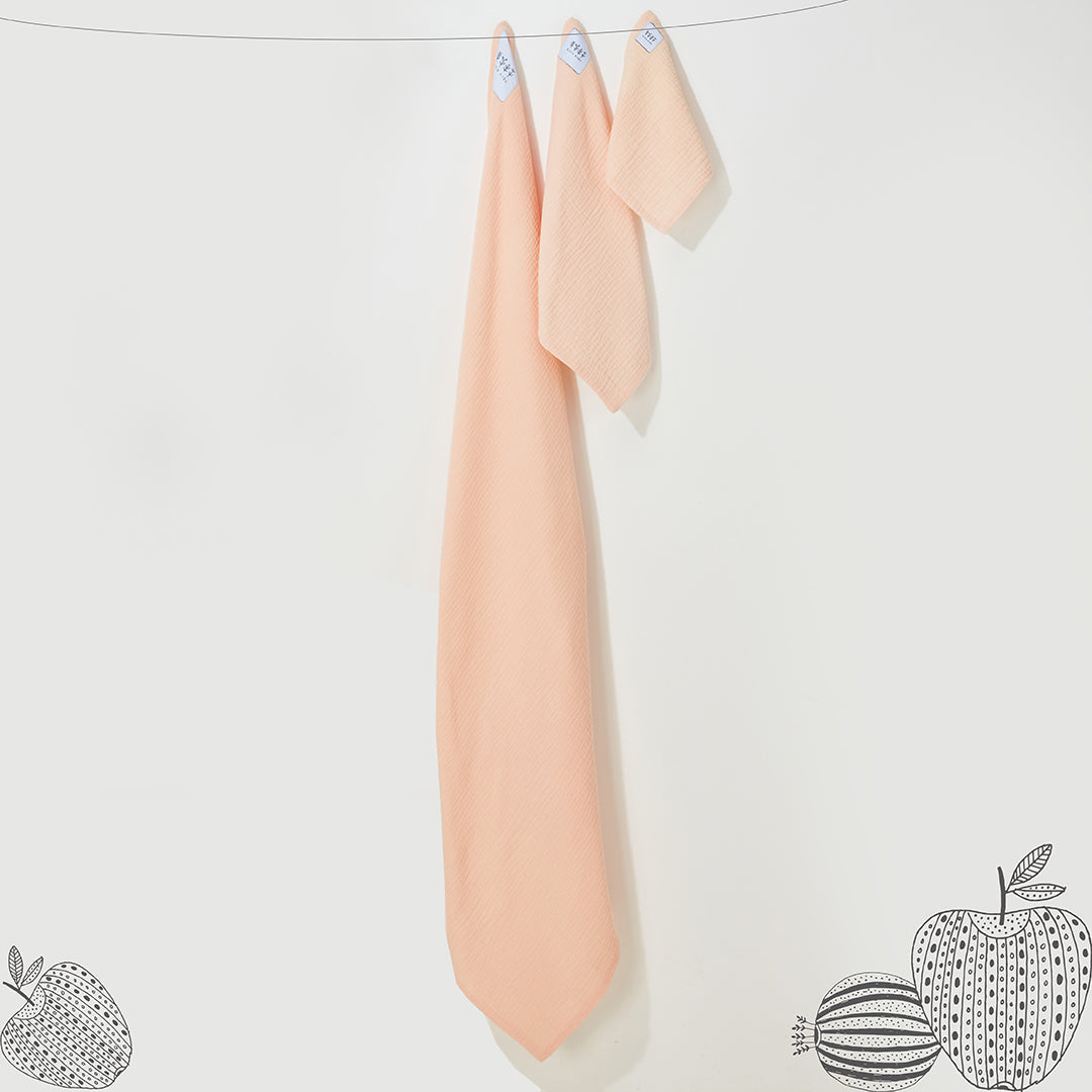 Trois langes en mousseline de coton couleur pêche de tailles différentes, suspendus sur un fil avec de légères illustrations de fruits au sol.