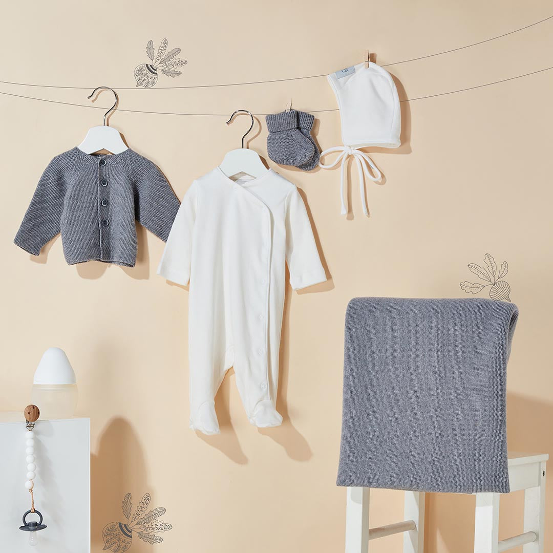 Ensemble de naissance suspendu comprenant un gilet en tricot gris, un pyjama blanc, des petits chaussons en laine, un bonnet et une couverture assortie.