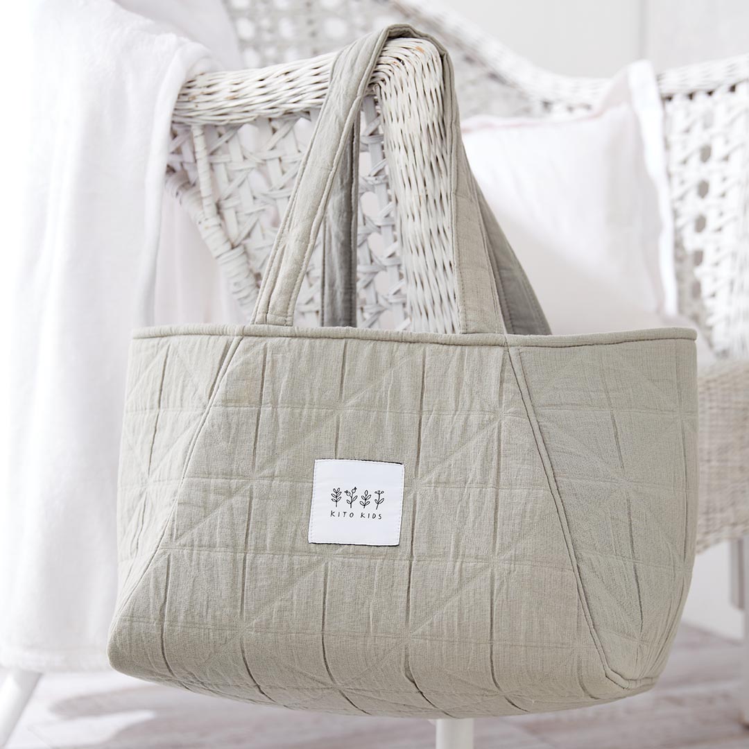 Sac de maternité matelassé beige suspendu au dossier d'un fauteuil en osier blanc dans une chambre de bébé.